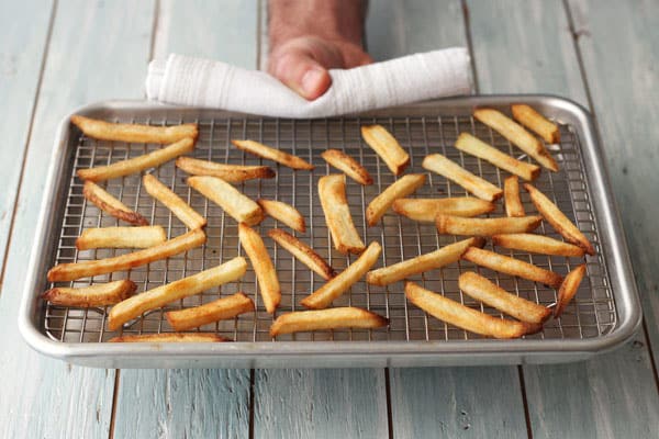 Hand holding a pan of cooked fries on a rack inside a sheet pan.