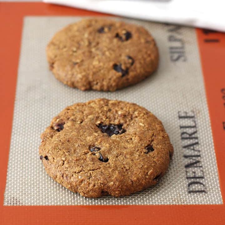 Two large cookies on a cookie sheet lined with a silicone baking mat.