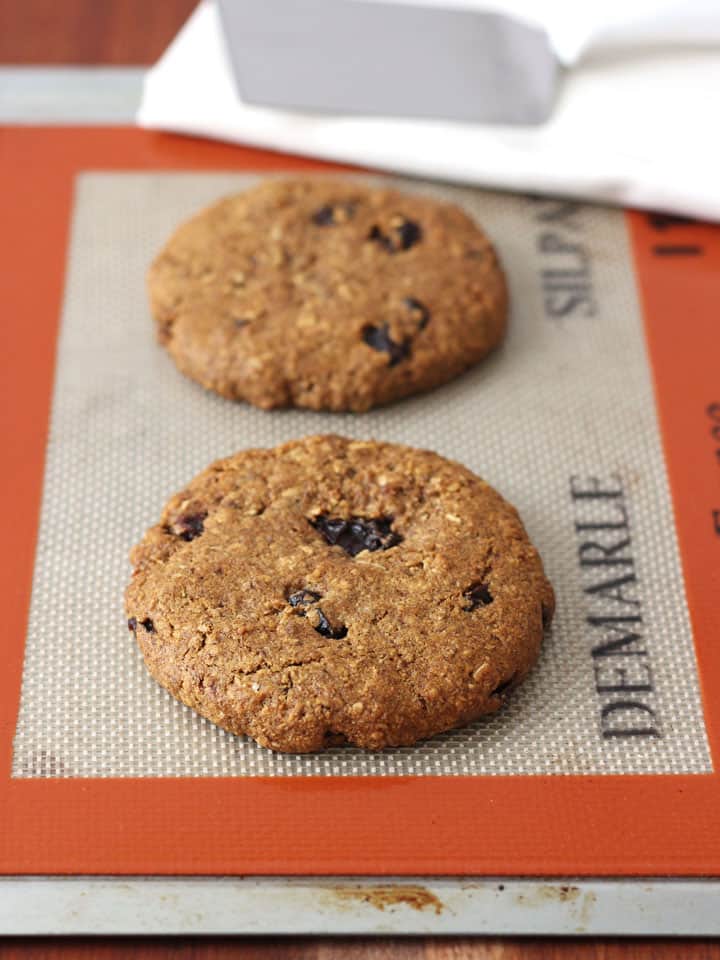 Two large cookies on a cookie sheet lined with a silicone baking mat.