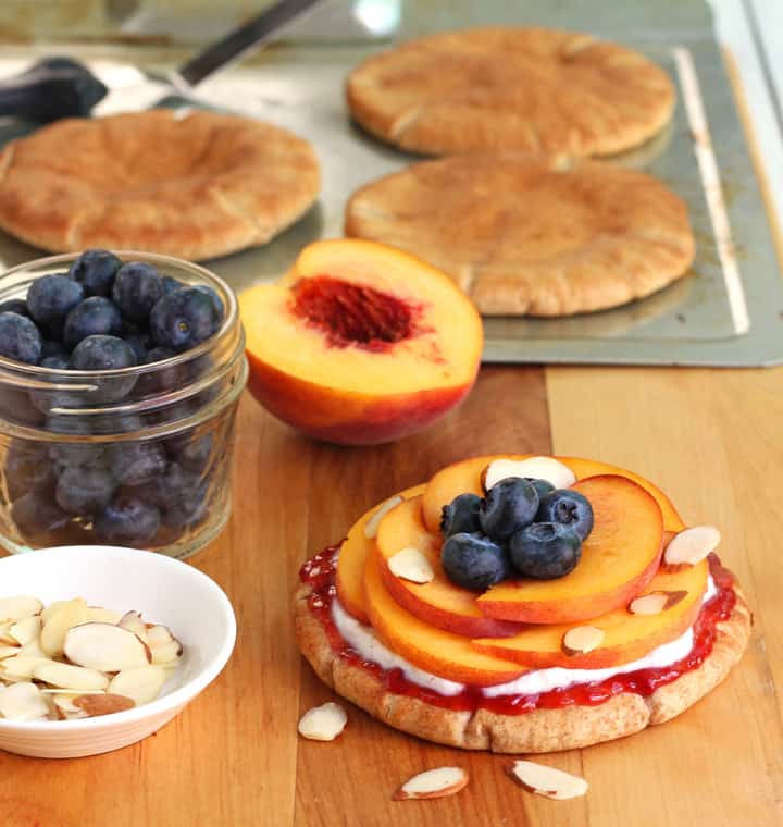 Cutting board with toasted pitas, blueberries, fresh peach, and almond slices.