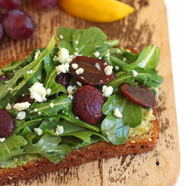 Toast topped with avocado, salad, and balsamic roasted grapes on a cutting board.