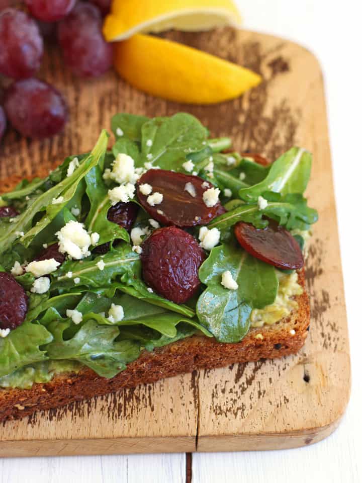 Toast topped with avocado, salad, and balsamic roasted grapes on a cutting board.