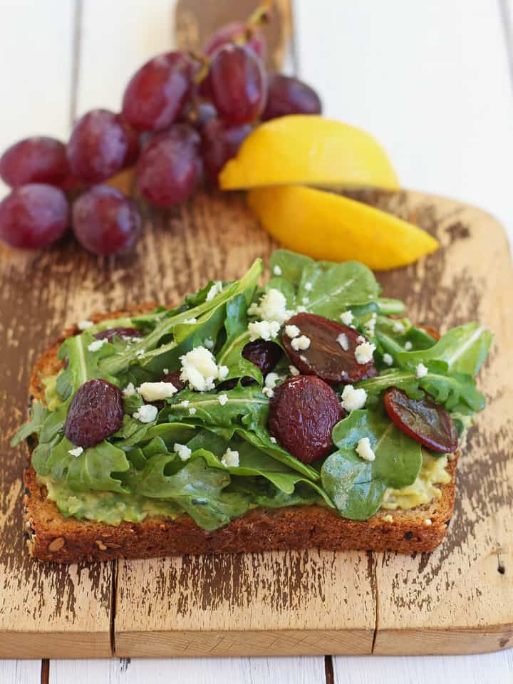 A small cutting board with avocado toast, grapes, and lemon wedges.