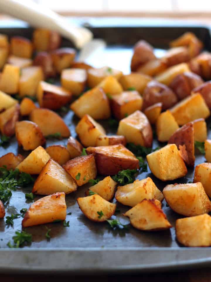 A dark-colored sheet pan with roasted potato cubes sprinkled with parsley.