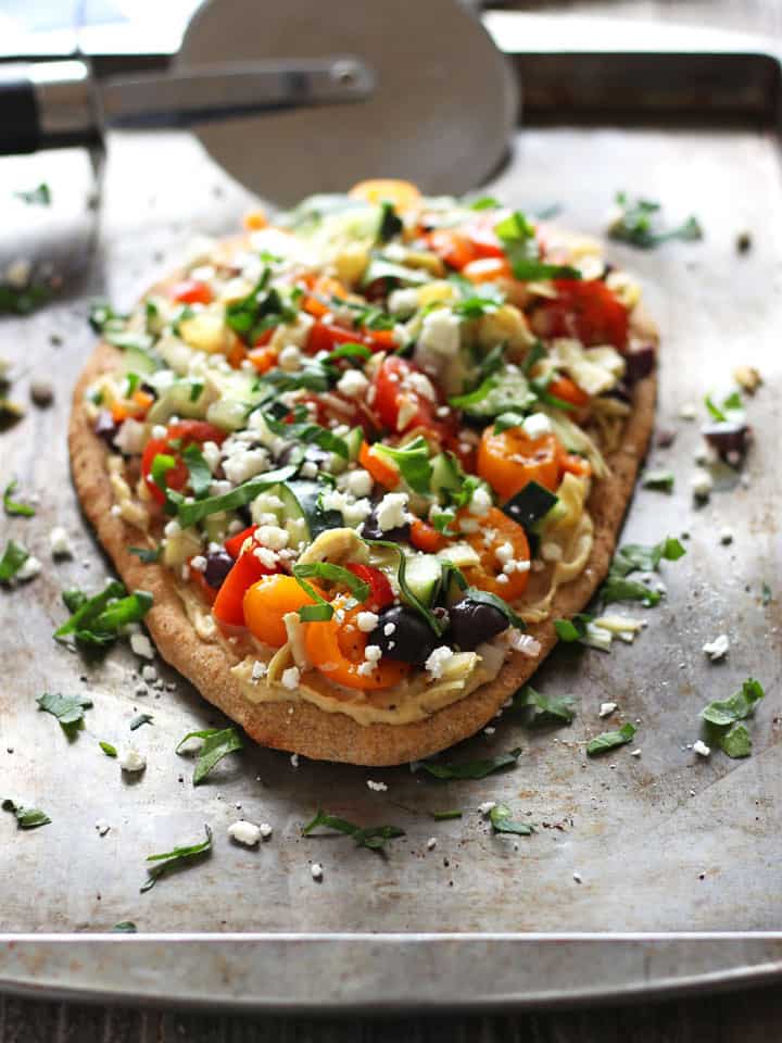 Vegetable topped flatbread on a sheet pan with a pizza cutter.