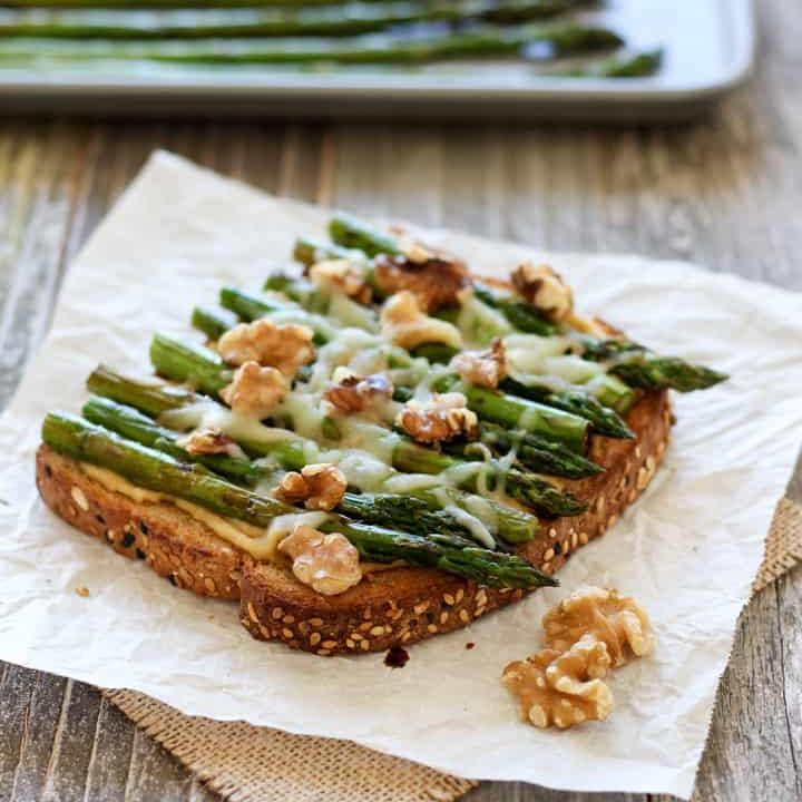 Piece of toast topped with hummus, asparagus, and walnuts.