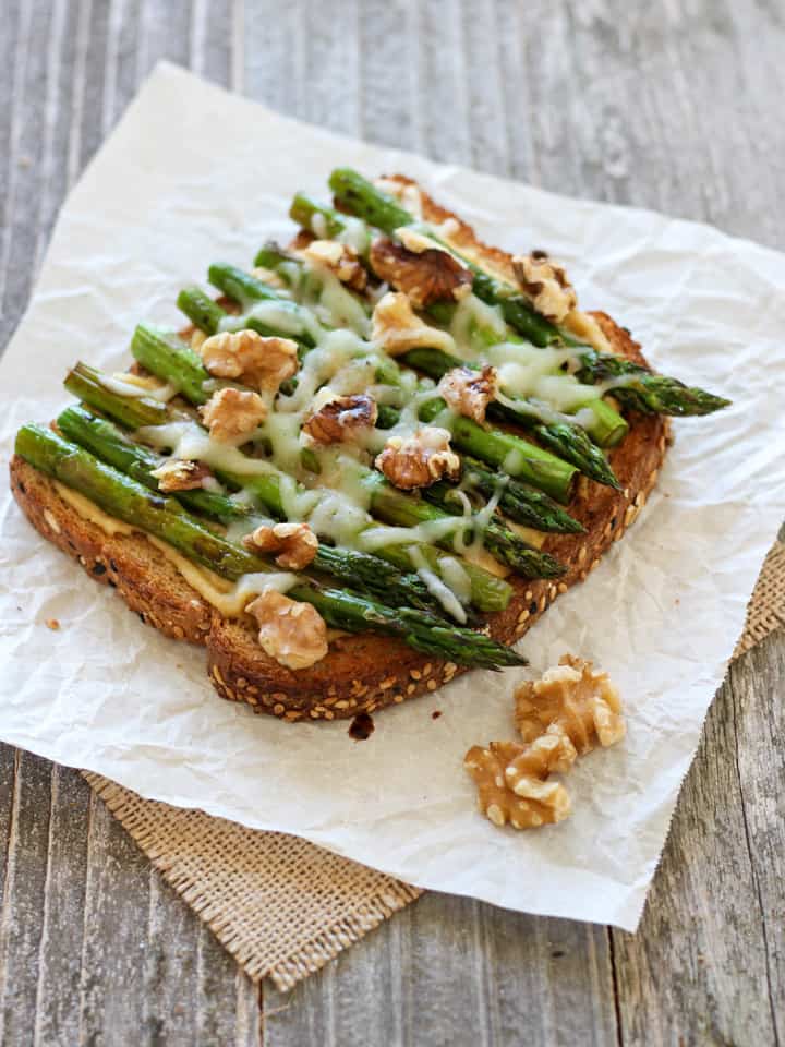 Piece of toasted bread topped with hummus, asparagus spears, and shredded cheese.