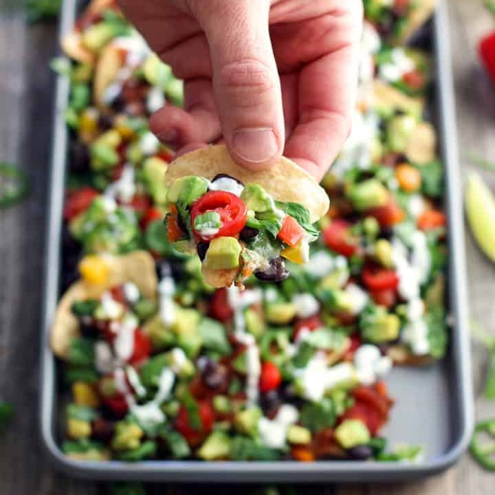 Hand scooping up a chip from a pan of veggie nachos.