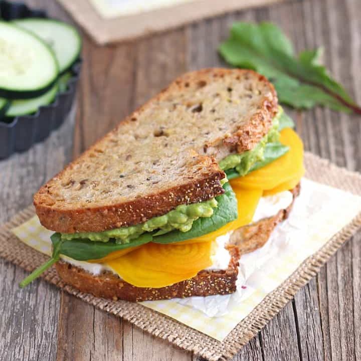 Beet and avocado sandwiches on a wooden table with a bowl of cucumber slices.