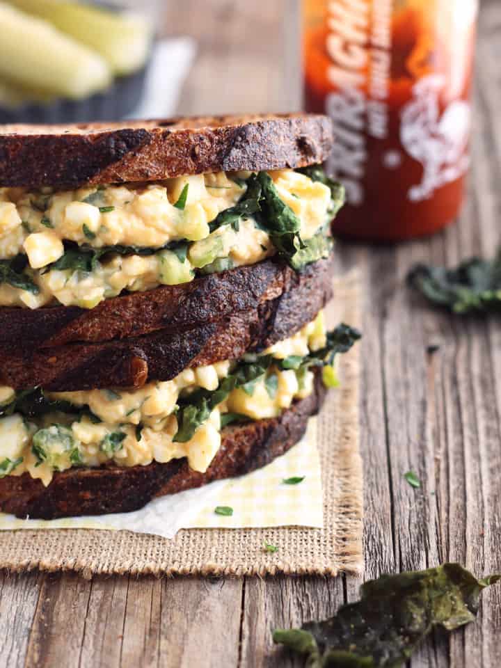 Stacked egg salad sandwiches next to a bottle of sriracha sauce.