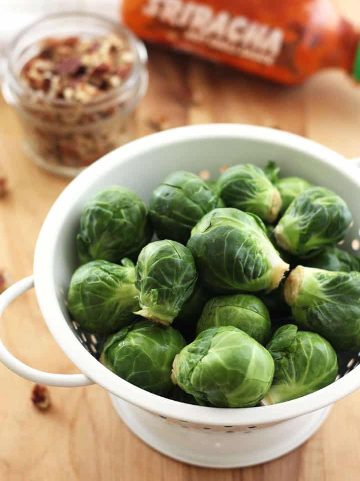 Fresh Brussels sprouts in a white colander on a cutting board.