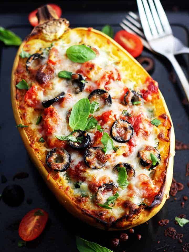 A cheesy spaghetti squash boat topped with olives and tomatoes on a dark roasting pan.