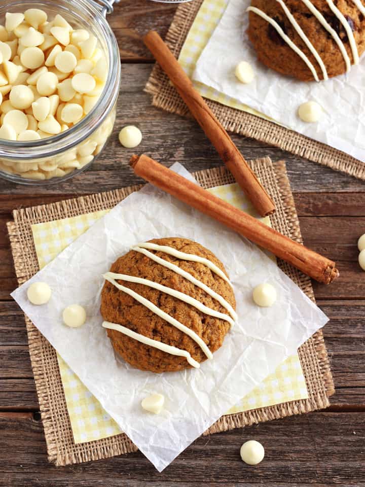 A cookie on a wood table with a jar of white chocolate chips and cinnamon sticks.