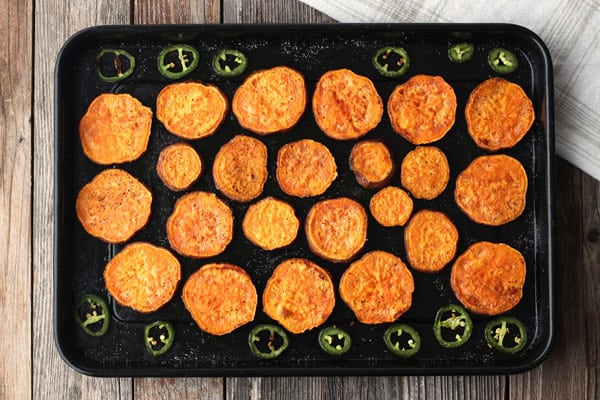 Baked sweet potato slices and jalapeno slices on a dark sheet pan.