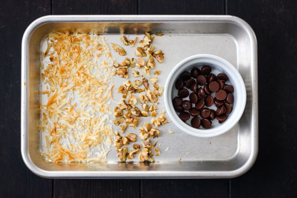 Toasted coconut and nuts on a baking sheet with melted chocolate in a ramekin.