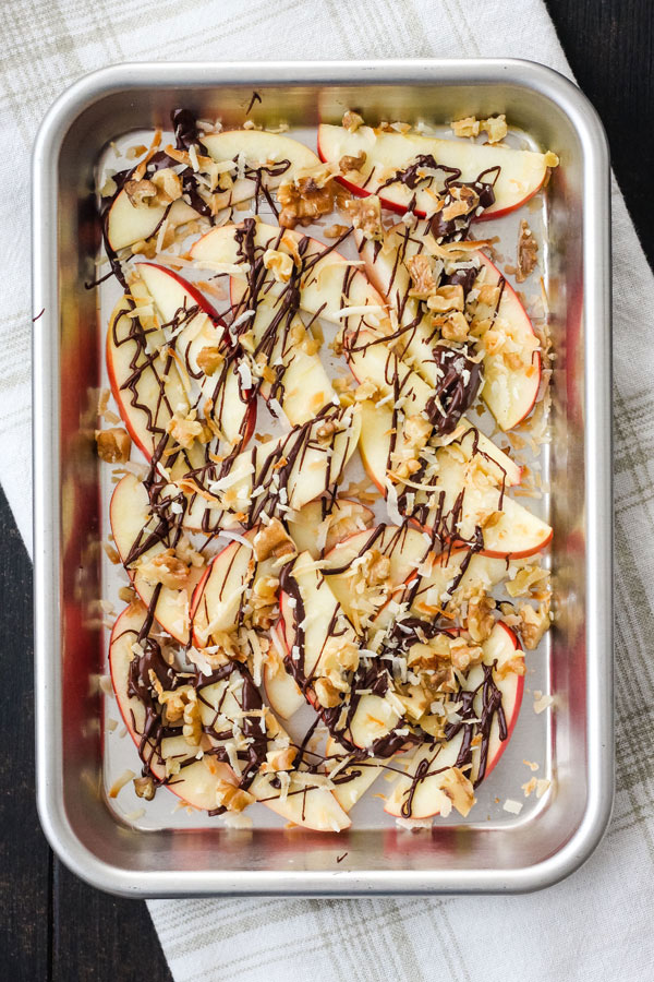 Apple nachos with drizzled chocolate and chopped nuts on a small baking sheet.