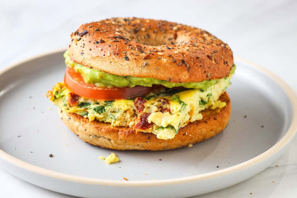 Bagel sandwich layered with eggs, tomato slices, and guacamole on a plate.