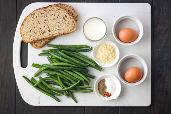 Two bread slices, green beans, eggs, cheese, and cream on a cutting board.