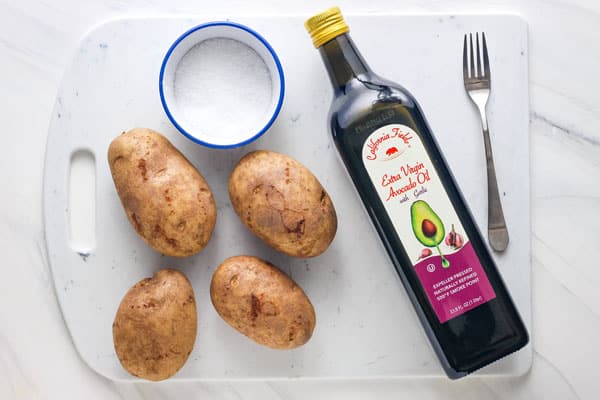 Russet potatoes, bottle of oil, and ramekin of salt on a cutting board.