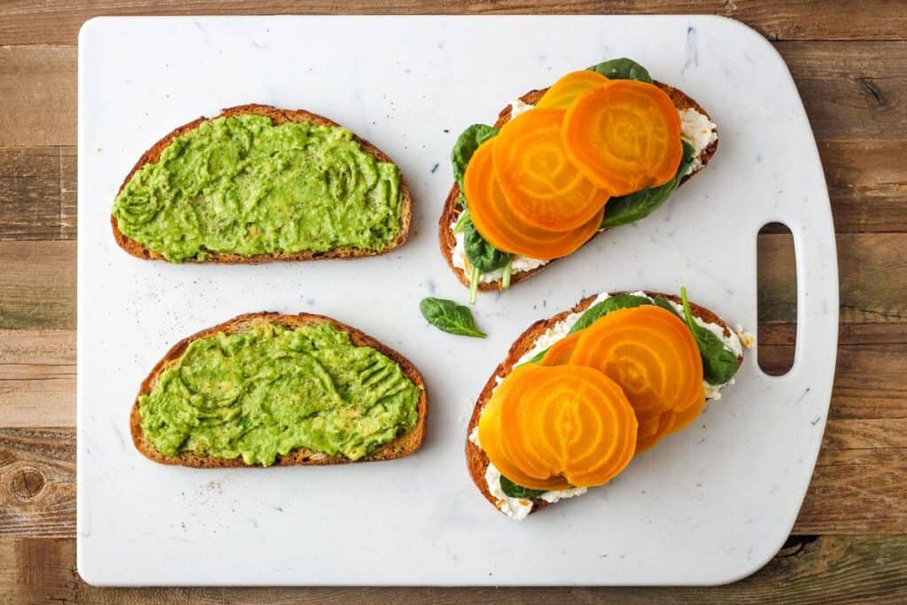 Slices of bread topped with avocado and sliced golden beets on cutting board.