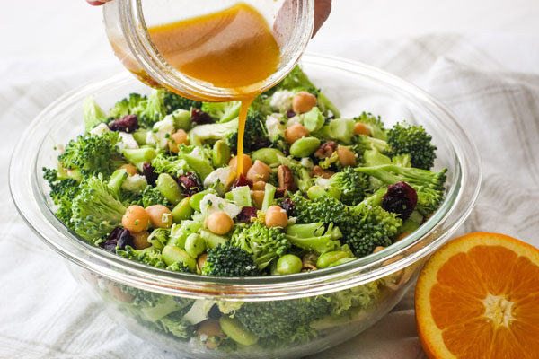 Hand pouring citrus vinaigrette from a mason jar over a bowl of broccoli salad. 