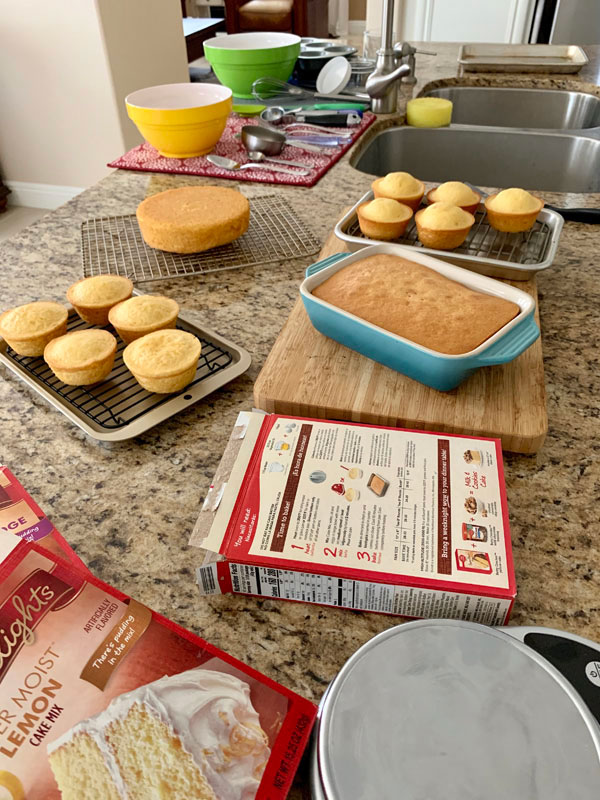 A kitchen counter with cake mix boxes, cupcakes, mini cakes, and dirty dishes.