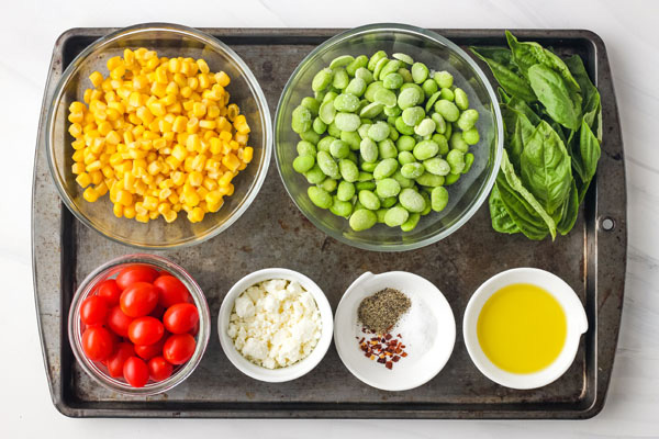 Bowls with corn, edamame, grape tomatoes, feta and basil on a baking sheet.