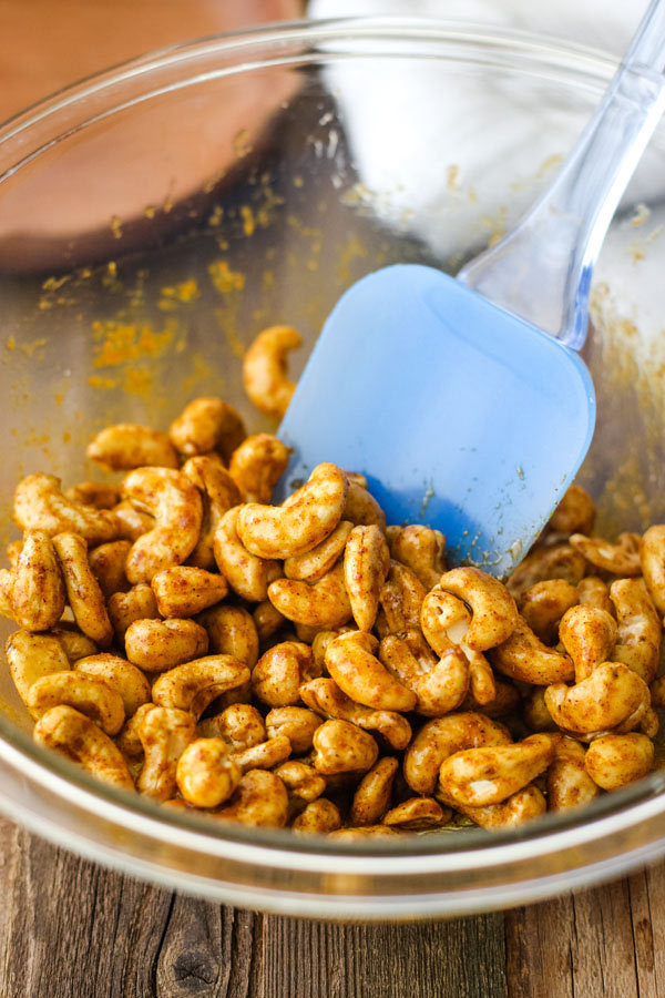 Cashews and seasonings in a glass bowl with a blue spatula.