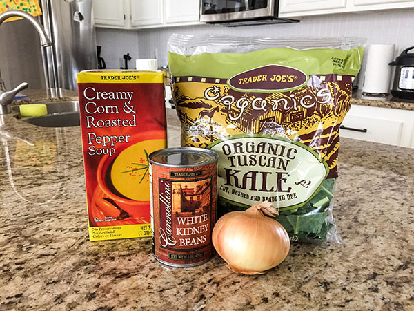 A box of soup, canned beans, bag of kale and an onion on a kitchen counter.