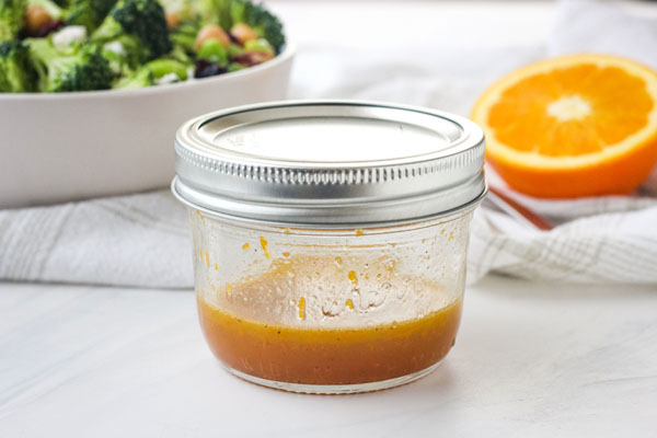 Dressing in a small mason jar with a bowl of salad and half an orange in the background.