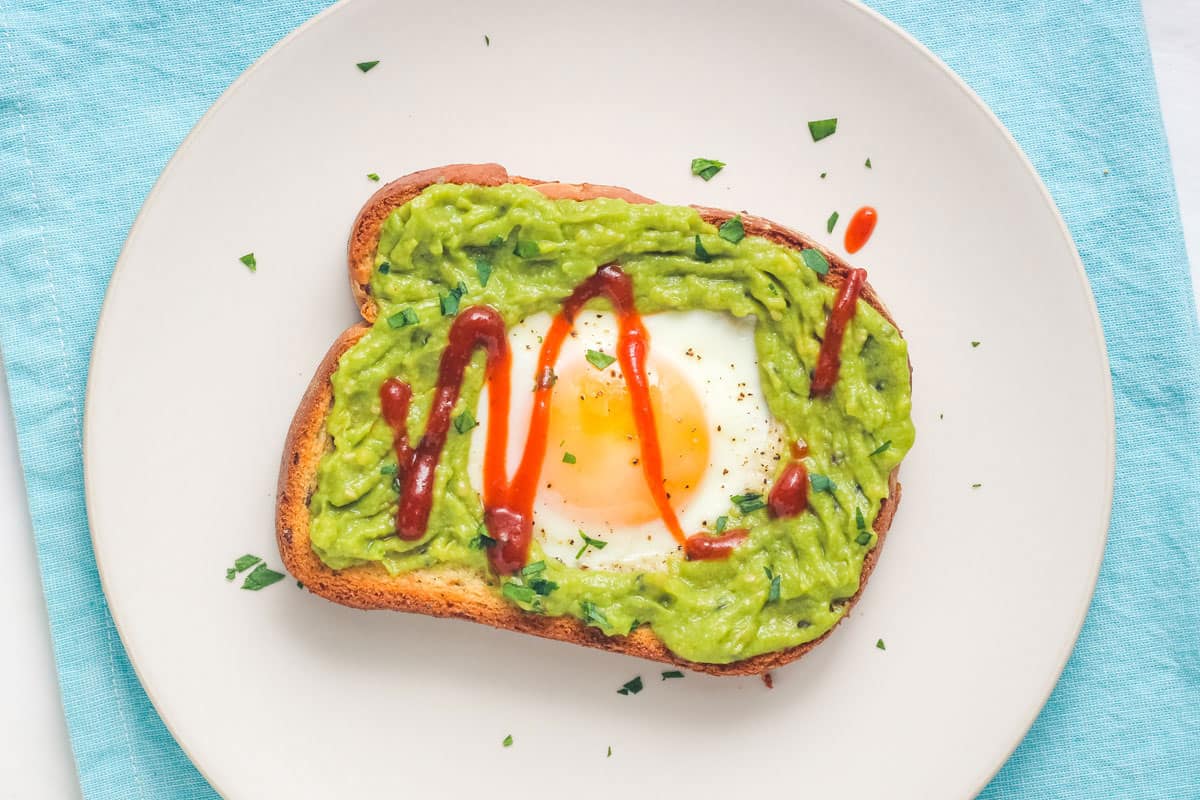 Overhead view of an egg cooked on a piece of toasted bread topped with avocado and hot sauce.