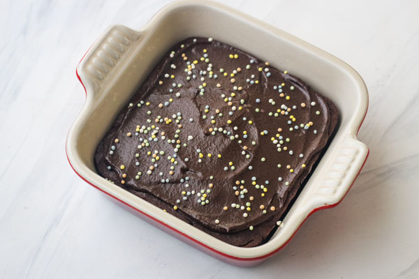 A mini chocolate cake with chocolate frosting and sprinkles in a 5x5 stoneware baking dish.