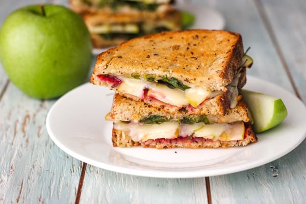 A white plate with two grilled cheese halves next to a green apple.