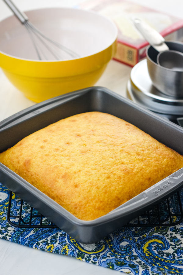 A yellow cake in an 8x8 baking pan, yellow mixing bowl and boxed cake mix on table.