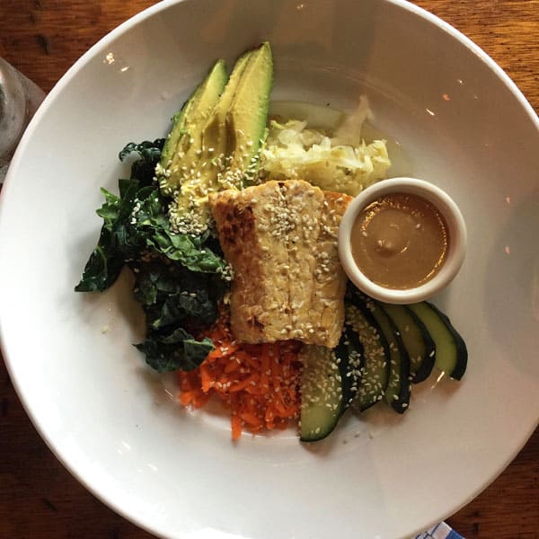 White bowl with tempeh, kale, sliced avocado, carrots and a brown dressing.