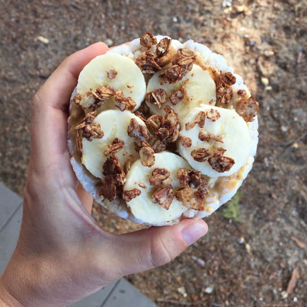 A hand holding a rice cake with peanut butter, banana slices and chocolate granola.