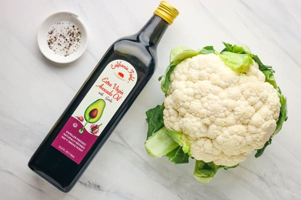 Large head of cauliflower, bottle of avocado oil, and bowl of salt and pepper.