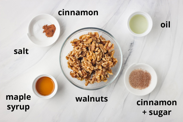 Bowls of walnuts, cinnamon, sugar, maple syrup, and oil on a white table.