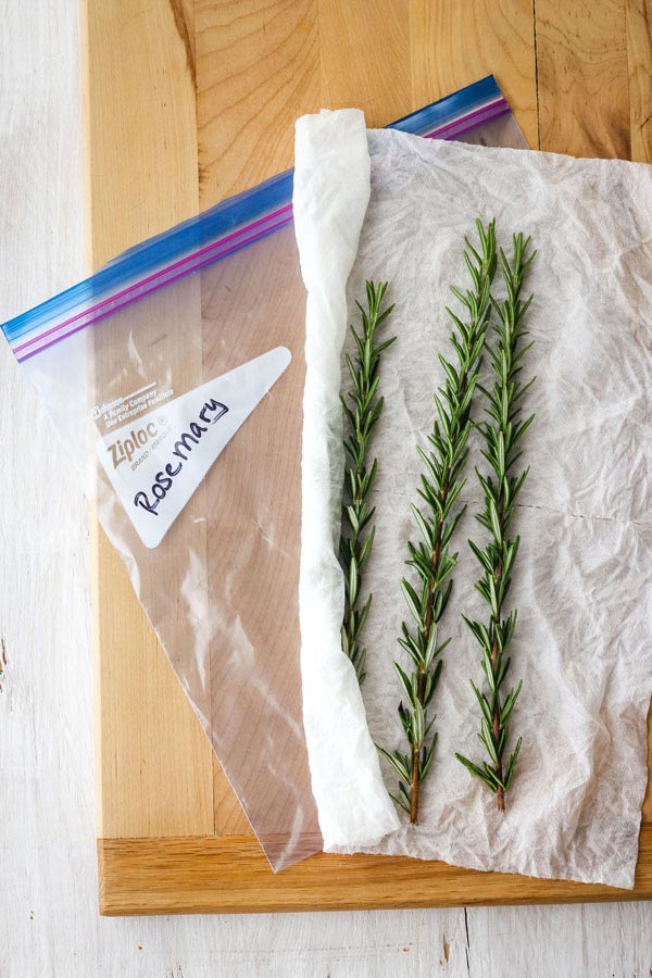 Rosemary on a damp paper towel next to a plastic bag with the words "rosemary" written on it.