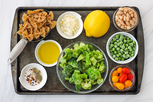 Dark metal baking sheet with bowls full of ingredients like frozen broccoli and pasta.