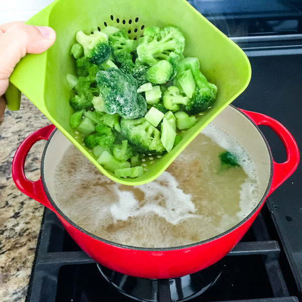 Hand holding a green colander with frozen broccoli held over a pot of boiling water.