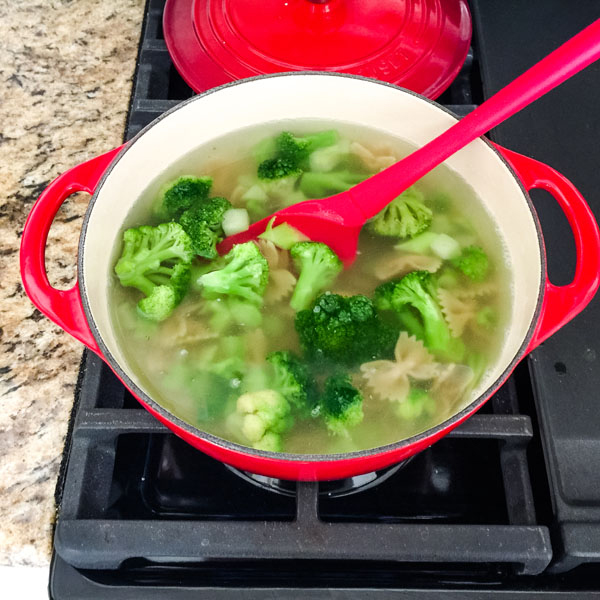 Red dutch oven full of boiling broccoli and pasta on a stovetop.