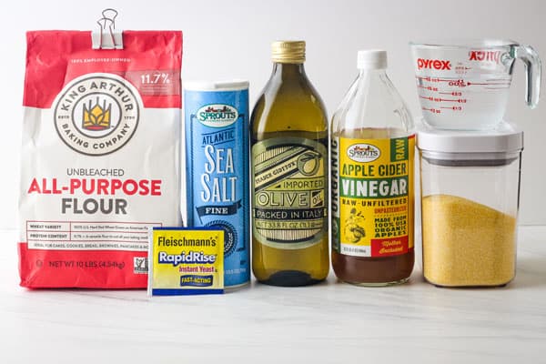 Bag of flour, yeast packet, jars of salt, oil, vinegar and cornmeal on a table.