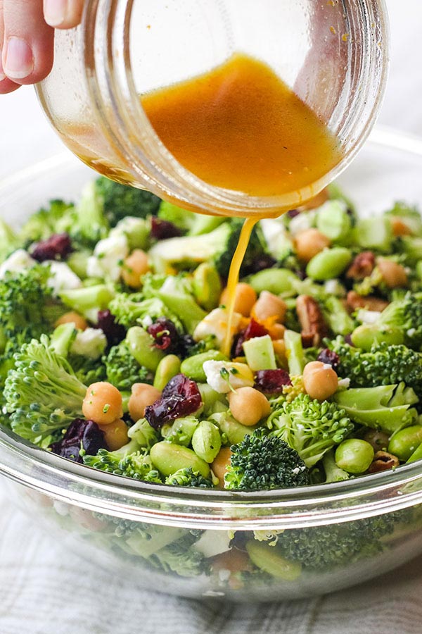 Hand pouring dressing from a mason jar over a broccoli salad.