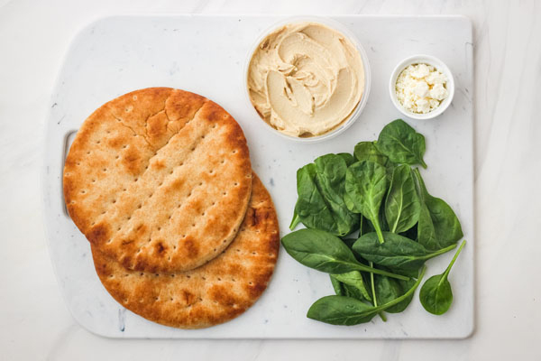 Pita breads, spinach, hummus and feta cheese on a cutting board.