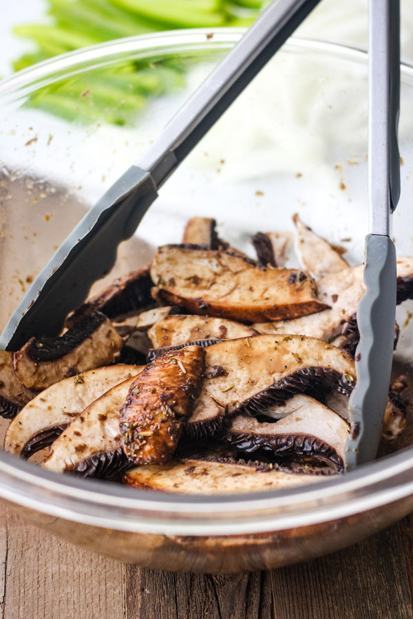 Sliced portobello mushrooms marinading in a glass bowl with tongs.