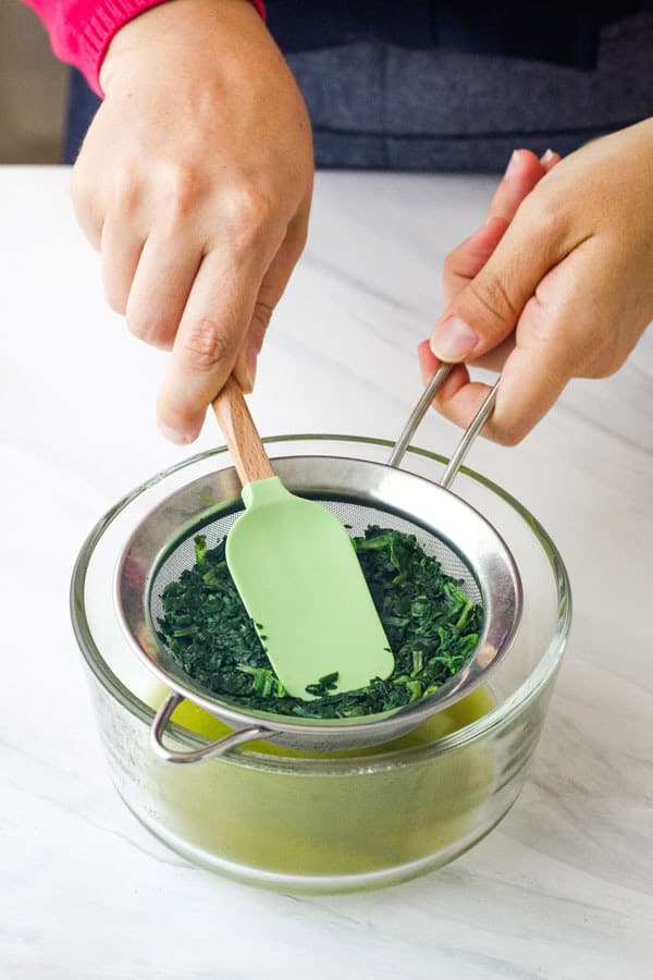 Green liquid in a glass bowl below collecting below spinach pressed in a colander.