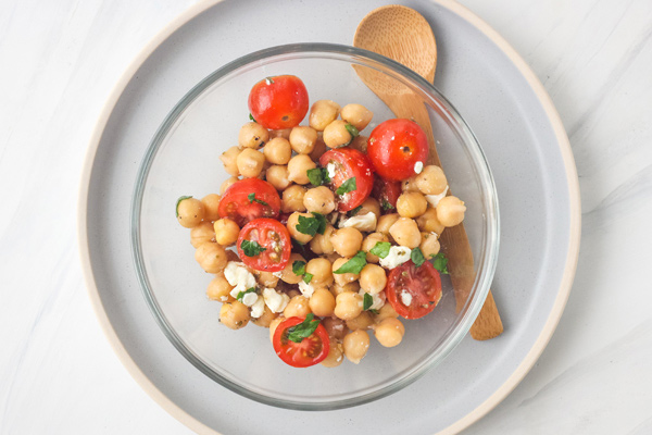 Chickpeas and tomatoes in a glass bowl next to a wooden spoon.