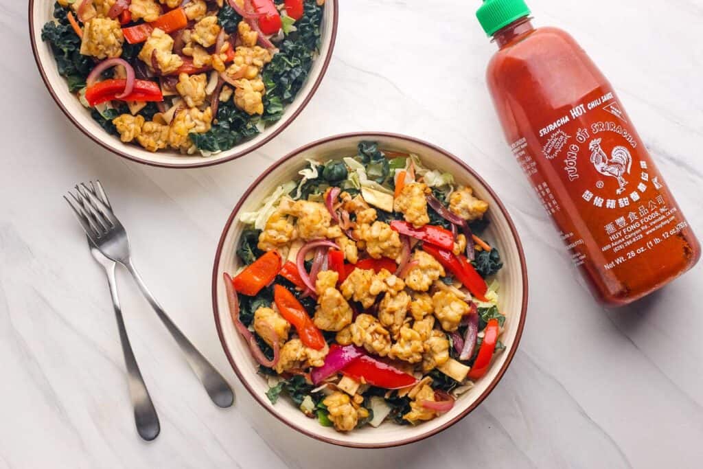 Overhead view of salad bowls with a large bottle of sriracha sauce.