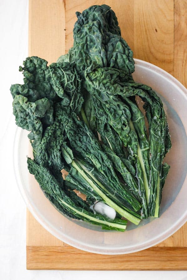 Overhead view of limp kale leaves in a bowl with water and ice cubes.